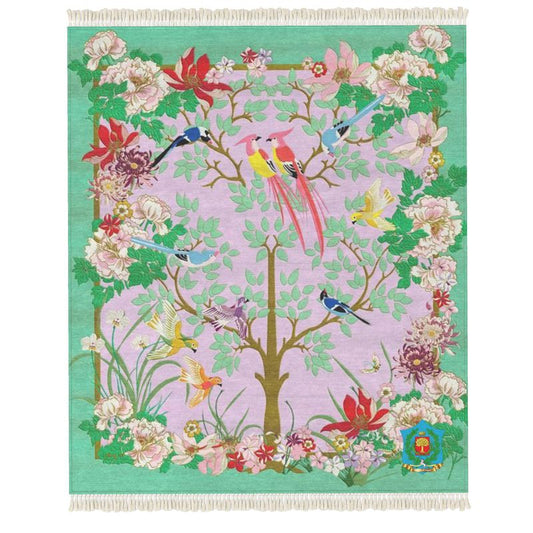 The Rug is a designer piece with a tapestry-like design featuring a central tree adorned with lush green leaves, surrounded by colorful birds and flowers. The pink and green backdrop highlights intricate floral patterns with birds in vibrant red, blue, yellow, and orange hues.