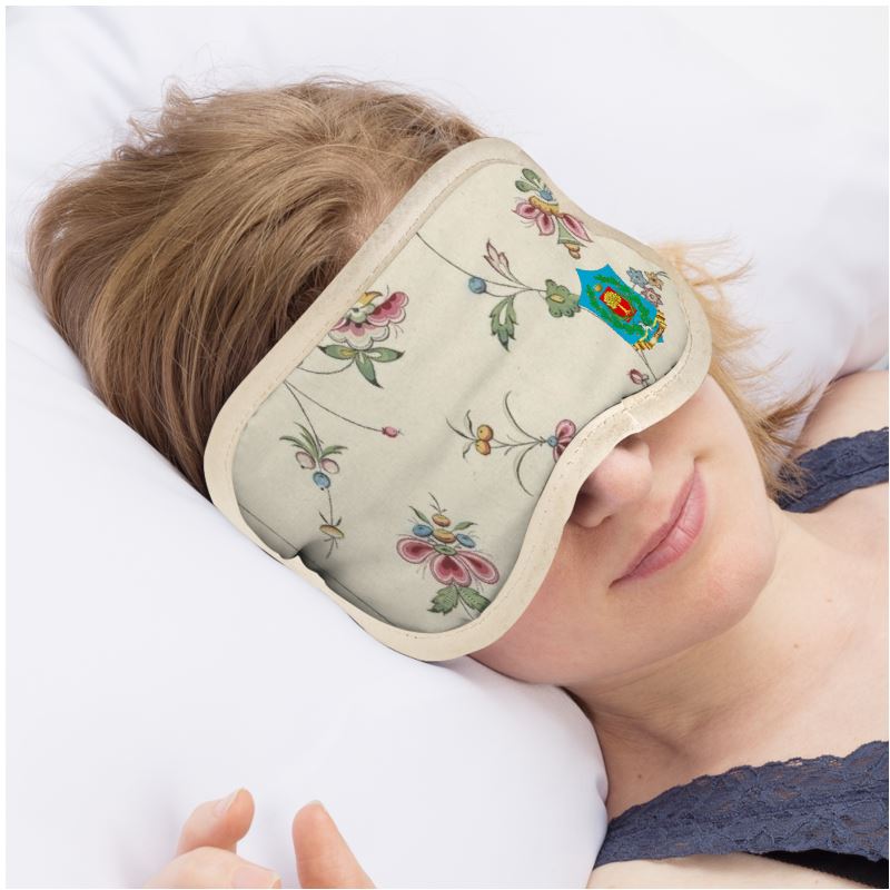 Lounging on white pillows, a person wears the FeelGood Sleep eye mask adorned with floral embroidery and a vibrant emblem. The light-haired individual, in a dark strap top, indulges in the luxury of soft satin fabrics for a restful slumber.